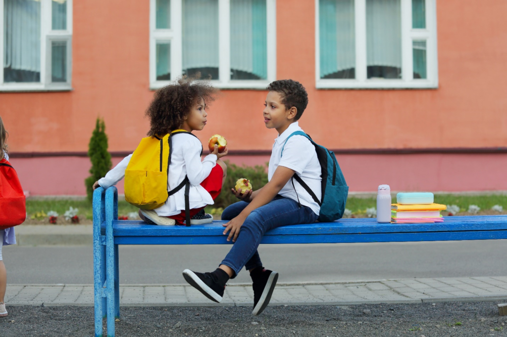 Two young students sitting on a bench eating apples.